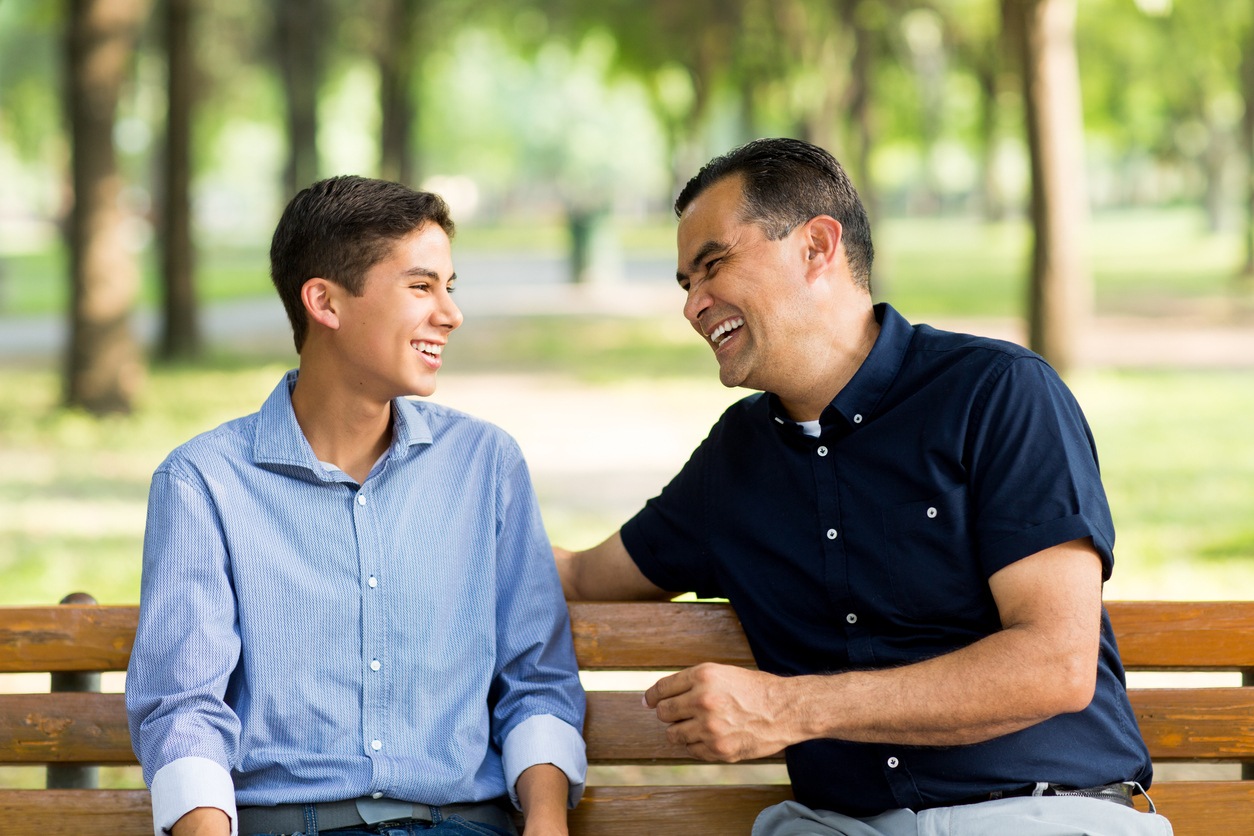 Hispanic father and son talking and laughing on a bench.