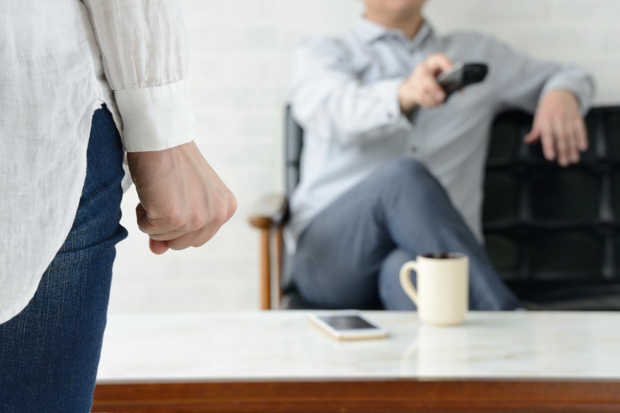 Woman with tight fist looking at man sitting on a couch using a remote.