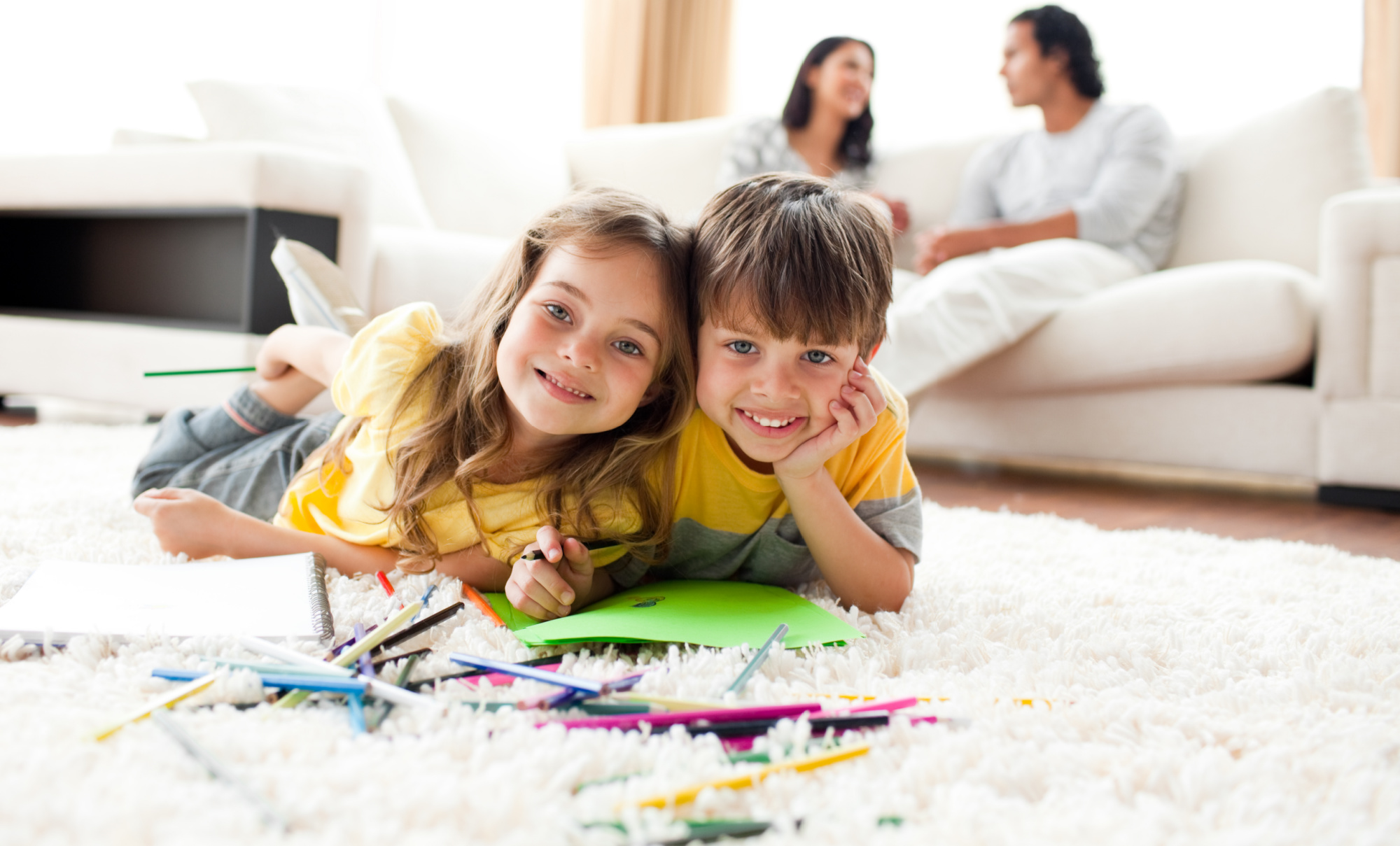 Young brother and sister playing together on the floor, parents in the background.